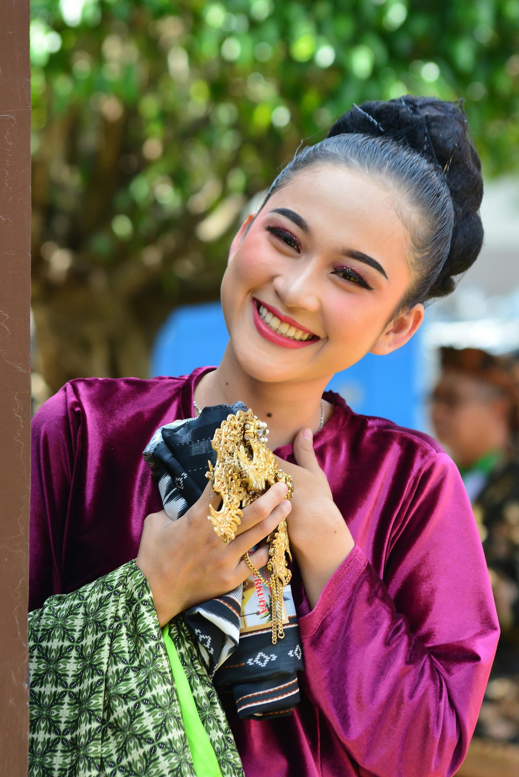 A woman dressed in colorful traditional Indonesian attire smiles brightly outdoors.