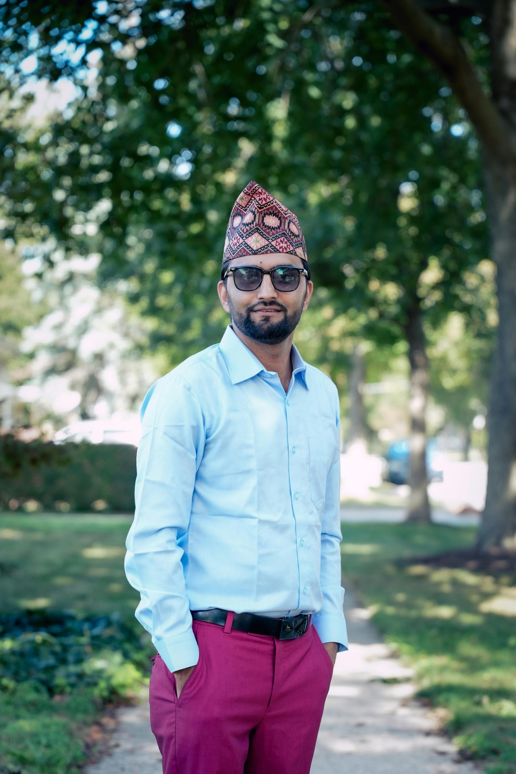 Confident man in traditional Nepali hat smiling outdoors, dressed stylishly in a serene park setting.